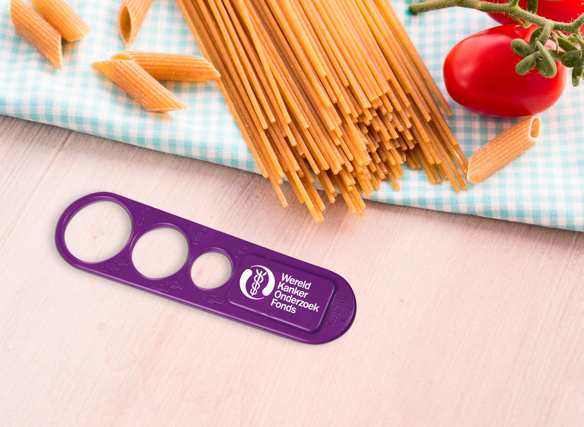 Paarse spaghettimeter met het logo van het Wereld Kanker Onderzoek Fonds in het wit. Op deze foto zien we de spaghettimeter op de keukentafel, naast ongekookte spaghetti.